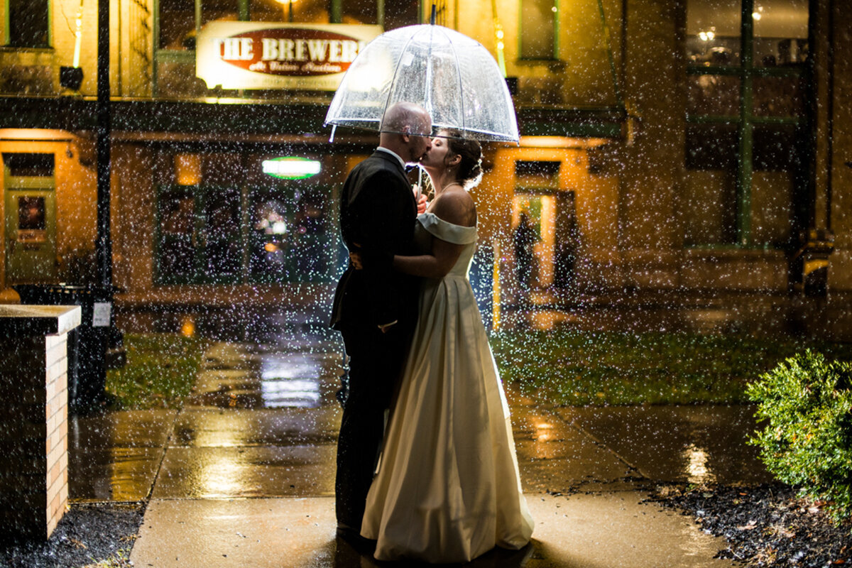 Bride and groom kissing under an umbrella in the rain at the the Brewerie at Union Station at night