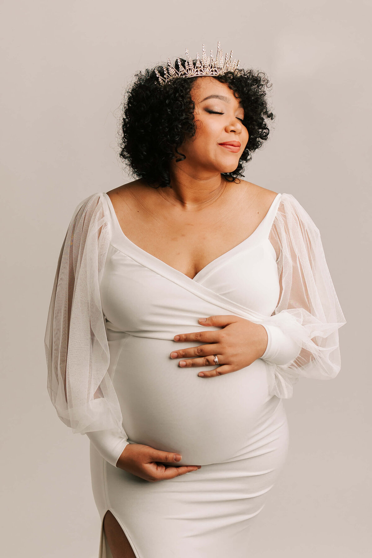 mom wearing crown wearing white dress smiling with eyes closed for maternity portrait