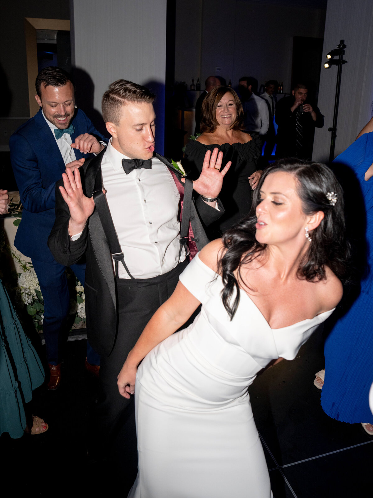 A couple dances energetically at a wedding reception. The woman in a white dress is in the foreground, while the man in a suit and bow tie dances behind her. Other guests are visible in the background, enjoying the festive atmosphere.