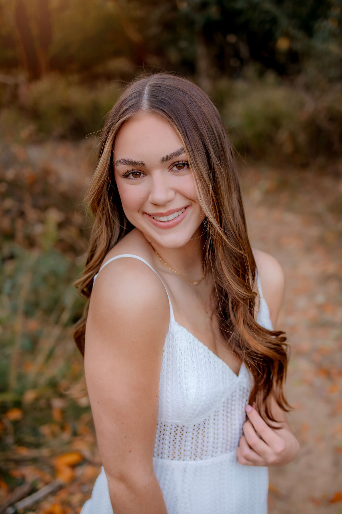 A very pretty young lady is wearing a white summer dress and standing next to a trail with wild flowers and posing for her senior photos with Foppiano Photography.