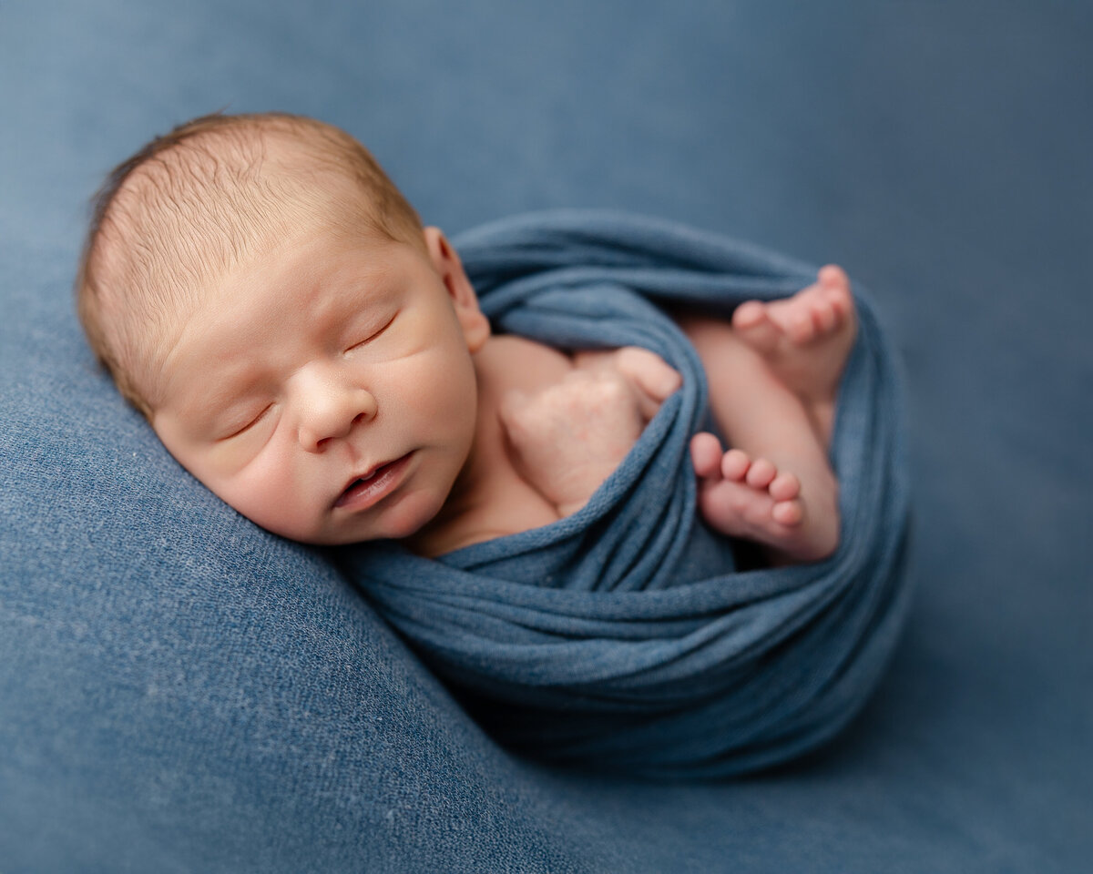Newborn baby boy wrapped in a soft blue swaddle, resting with one foot peeking out on a matching backdrop during his studio session. Captured by Inland Empire newborn photographer Kaitlyn Dawn Photography.