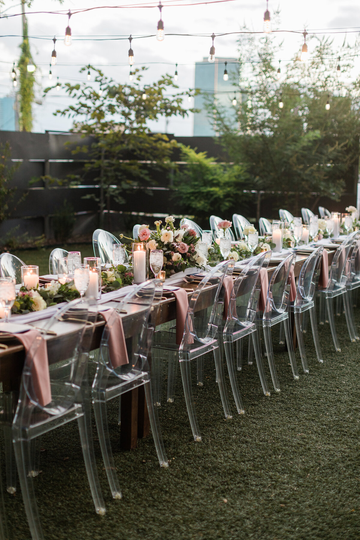 A detail shot of the main reception table at a wedding at Artspace111 in Fort Worth, Texas. The table is covered with fancy place settings, pale pink napkins, candles, and large array of floral centerpieces. The chairs are clear and surround both sides of the table.