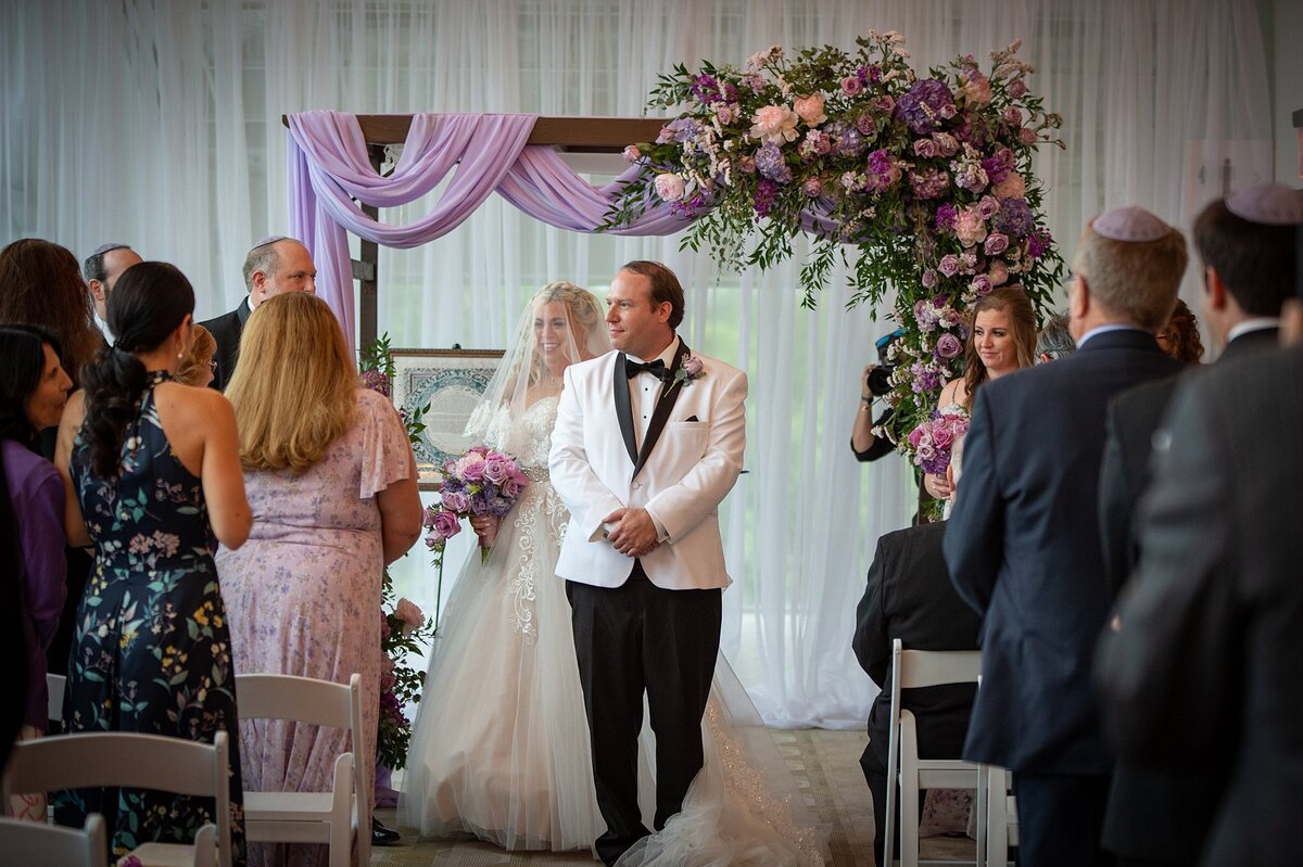 The bride wearing a veil over her face and a strapless lace wedding gown holding a bouquet of purple roses, lilac rose, pink roses and ivory hydrangea walks the seven circles around the groom who is wearing a white tuxedo jacket with a black shawl collar and black pants as they stand in front of their guests under the chuppah at Noah Liff Opera Center. The wood chuppah is draped with light purple fabric and an extra large floral spray of purple roses, pink roses, light purple roses and purple flowers with greenery at The Liff Center in Nashville.