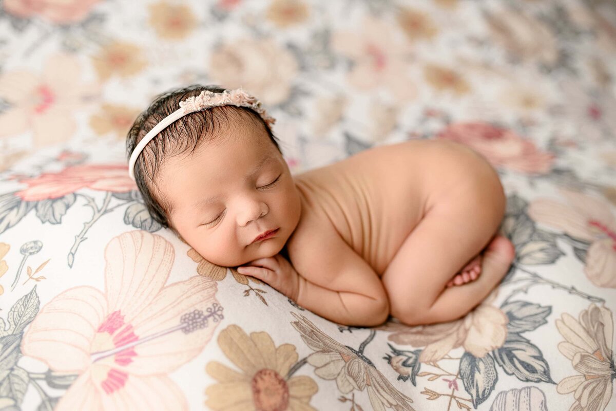 Newborn baby girl is naked and posed with a headband on a flower background all curled up gently during her Studio newborn session