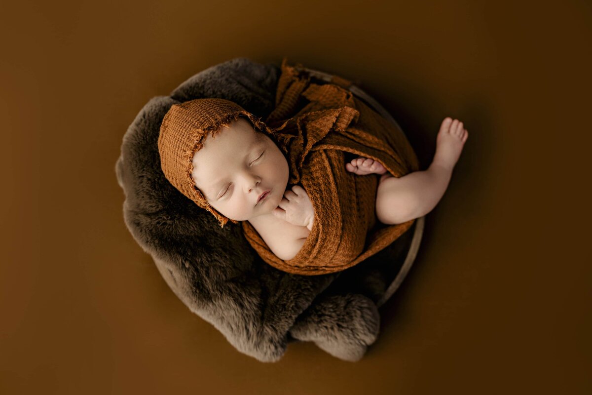 newborn from monroe wrapped in a rust colored wrap sits in a bowl with a matching studio background