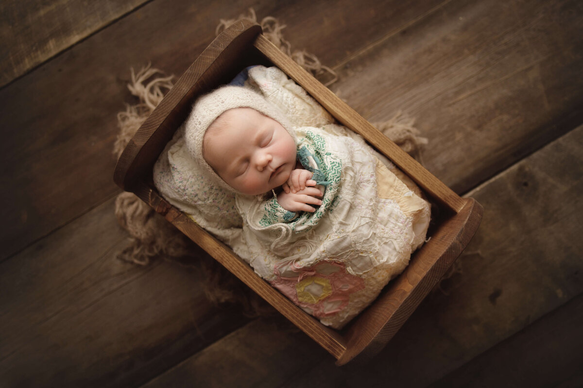 newborn baby in cradle wrapped in neutral quilt on wooden backdrop