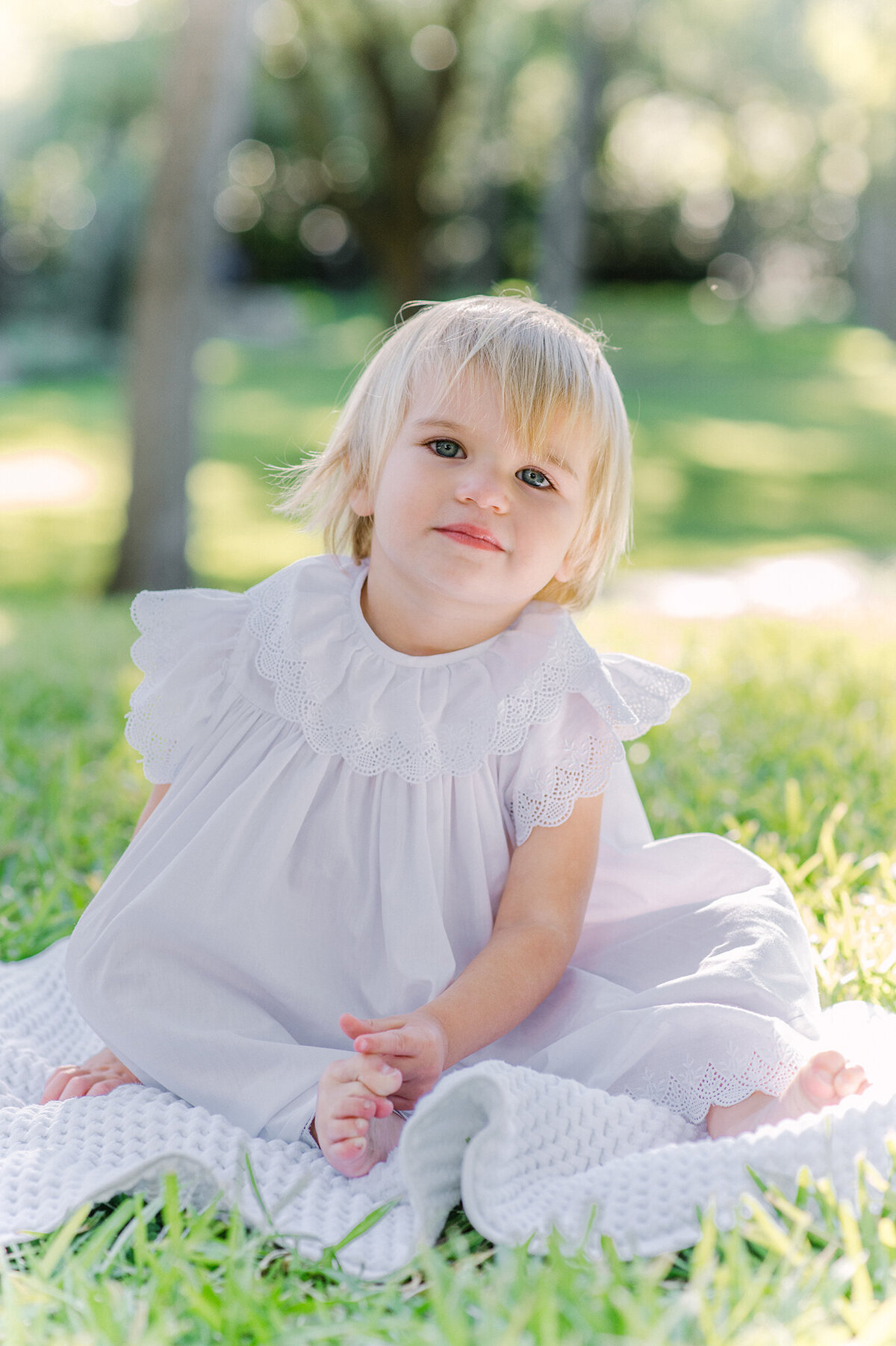 Blonde toddler sitting on a white blanket wearing a white dress in a park.