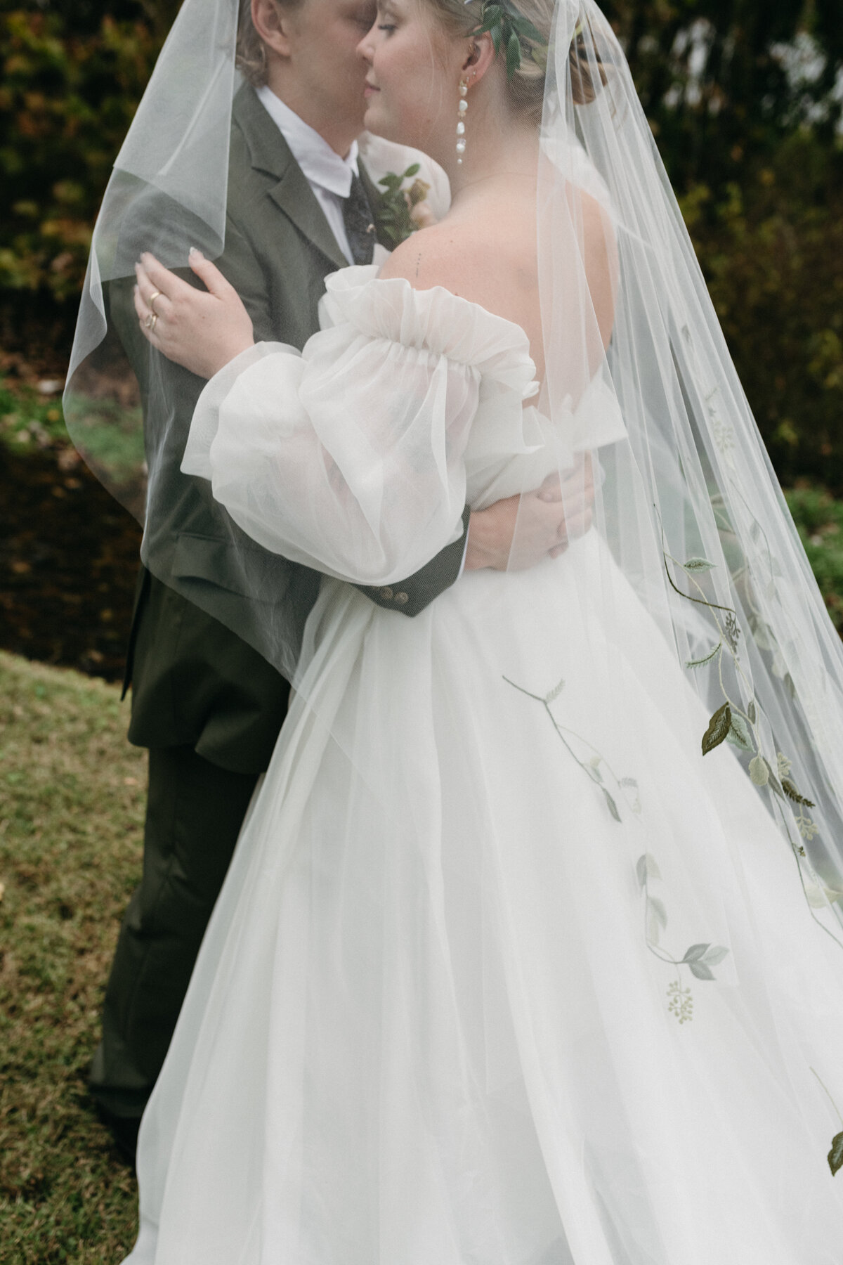 Romantic close-up of the bride’s embroidered tulle veil, puff-sleeve gown, and the groom’s arms around her, highlighting the soft, ethereal textures and garden-inspired floral details of their Northwest Arkansas wedding.