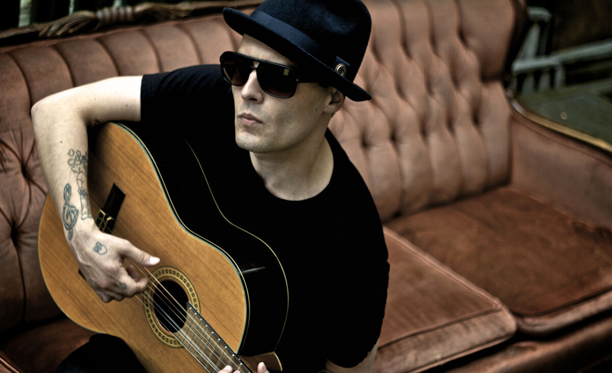 Male musician portrait G wearing black fedora black t shirt sunglasses sitting against old pink sofa playing guitar
