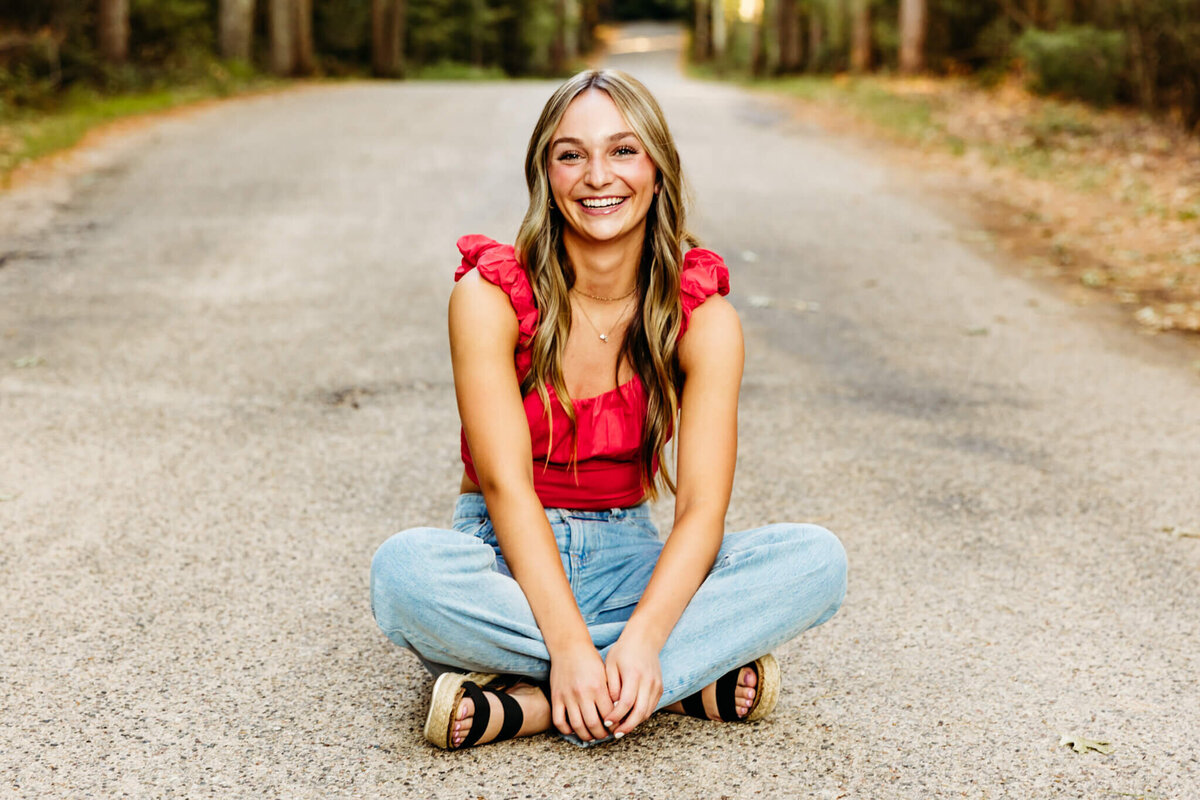 young lady in red top sitting crossed legged on a backroad for her senior photo session