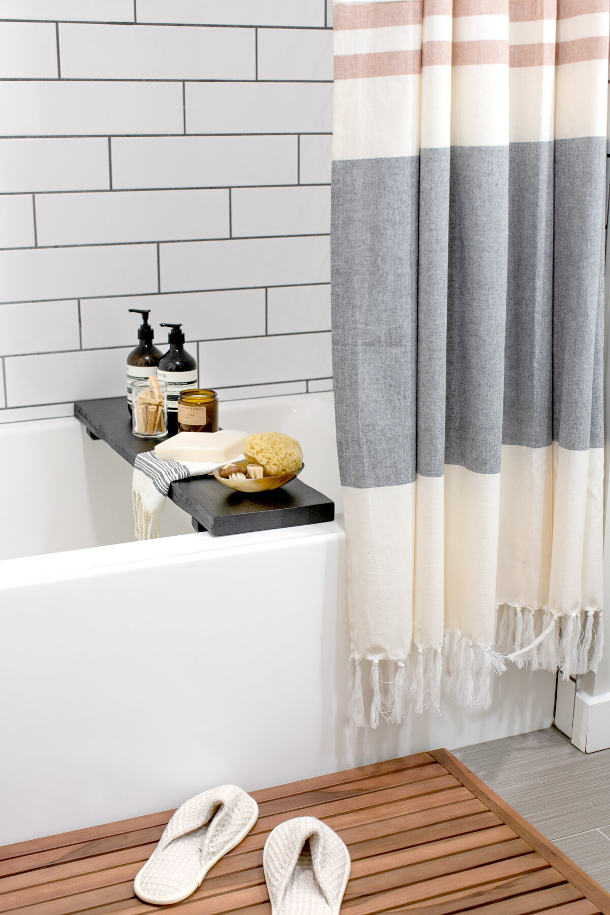 Bathroom with white bathtub, wooden bathmat with cotton slippers on top. Turkish style striped shower curtain covering white subway tiled wall. Black wooden tray with soaps and objects on top of tub