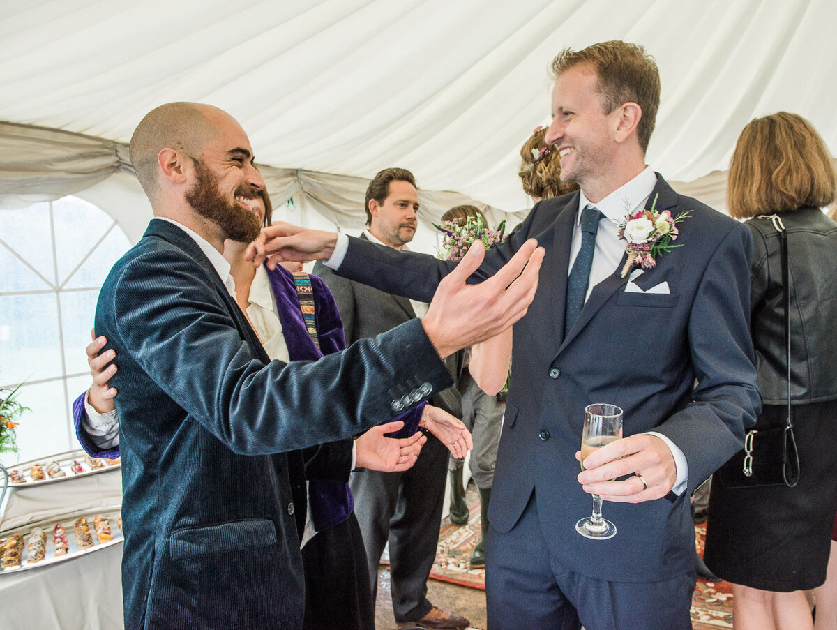 male guest in navy, velvet jacket embracing a groom in a navy wedding suit and wild flower buttonhole