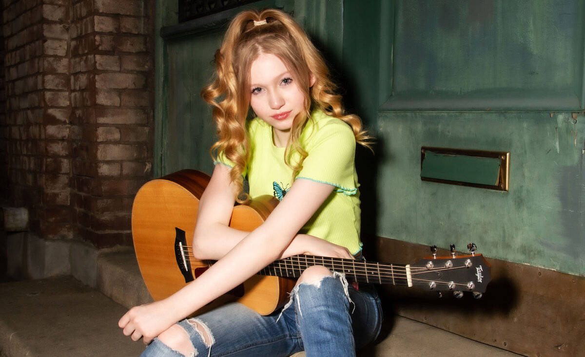 Female musician portrait Abigail Lewis wearing lime green shirt sitting with guitar against green door