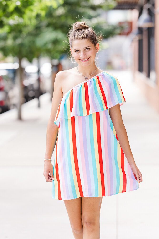 Pretty senior girl in a colorful over the shoulder mini dress walking by downtown Raleigh.