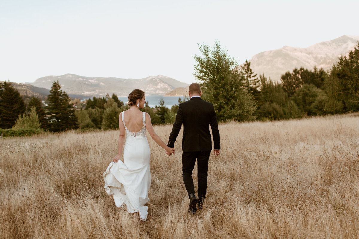 A bride and groom walk through a field at Skamania Lodge with a PNW mountain backdrop. Captured by Fort Worth wedding photographer, Megan Christine Studio
