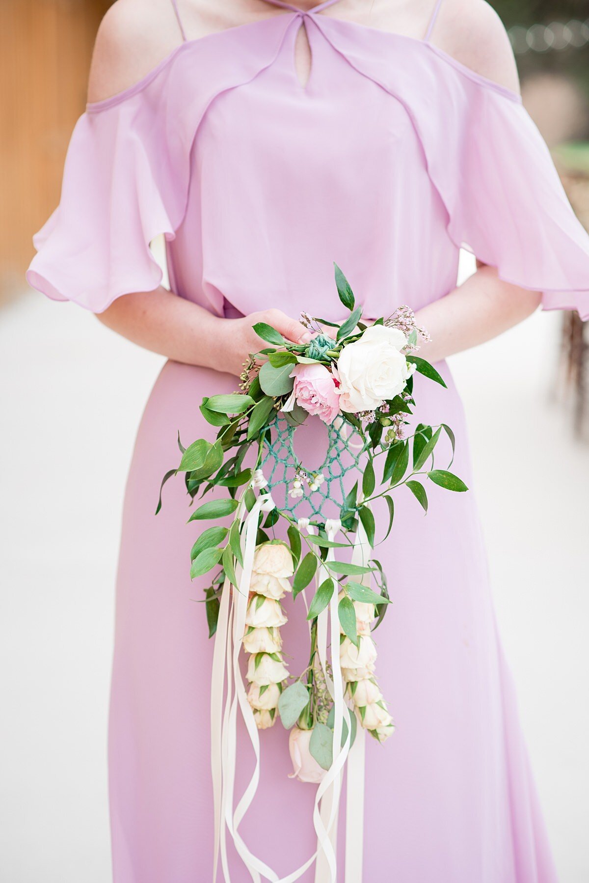 The bride is wearing a very light purple dress with a cold shoulder top accented by a large ruffle. Her bouquet is a large halo dreamcatcher accented by greenery, a white and pink flower and long streaming ivory ribbons.
