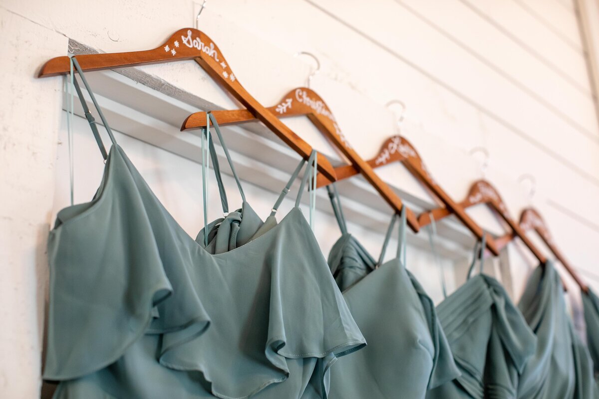 sage green bridesmaids dresses hang on wooden hangers with names at Morgan Creek Barn in Dripping Springs Texas