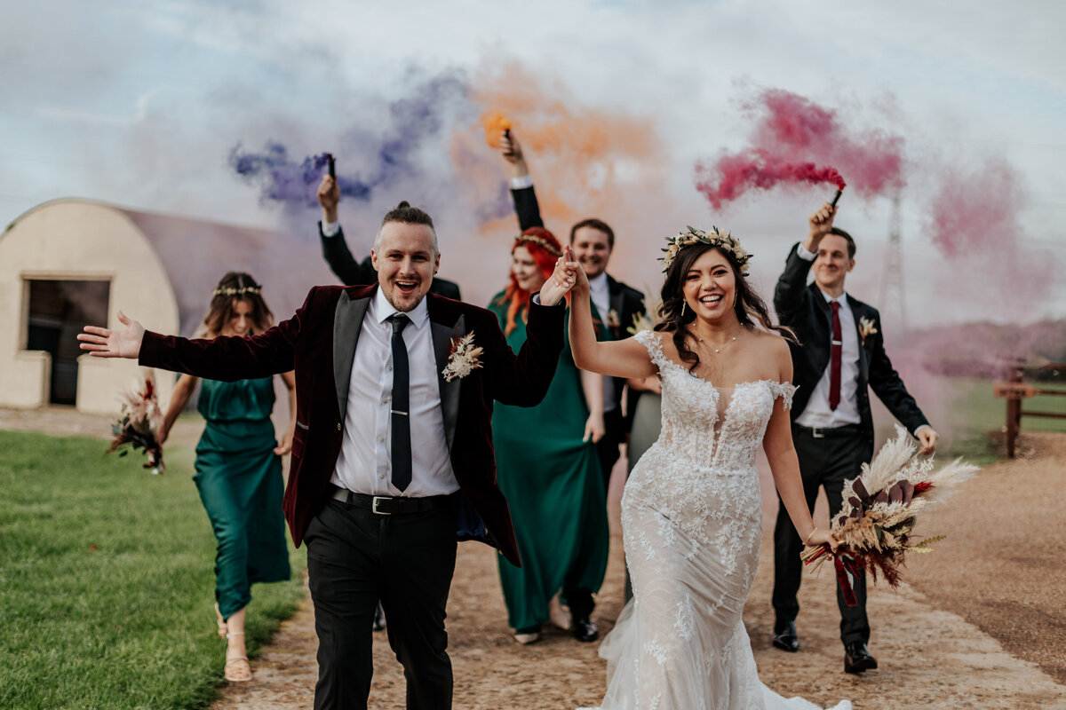 A bride and groom hold hands and walk infront while their wedding squad hold smoke flares in the background