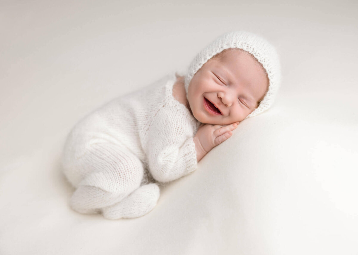 smiling newborn baby asleep on white fabric wearing a white knitted romper and bonnet