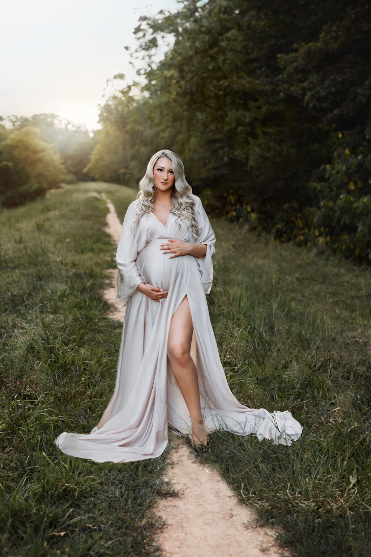 pregnant woman wearing a silky flowy sand colored maternity gown standing in a field with the sun setting behind her in a  field at her northern va maternity photo session