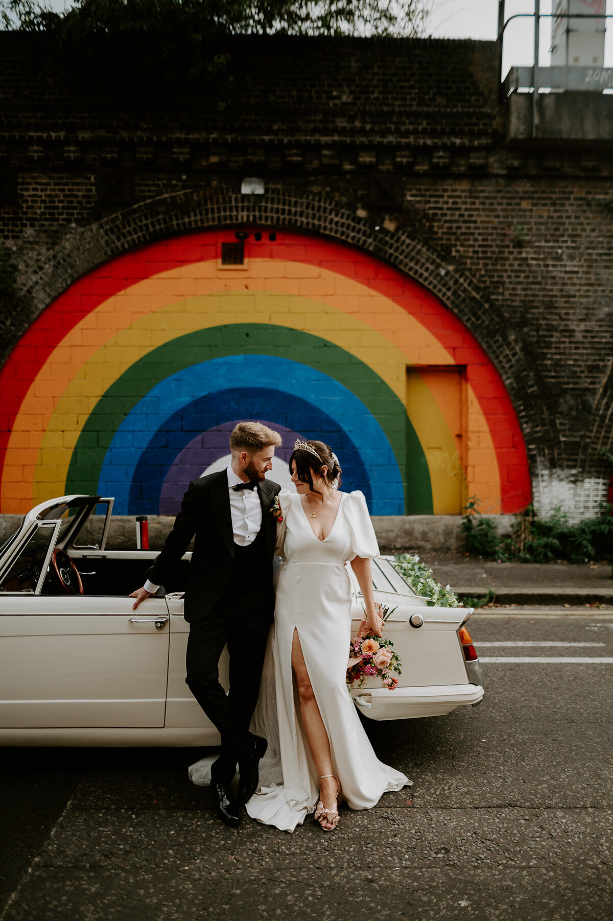 A bride and groom stand next to a Triumph convertable outside the rainbow mural at 100 Barrington on their wedding day.