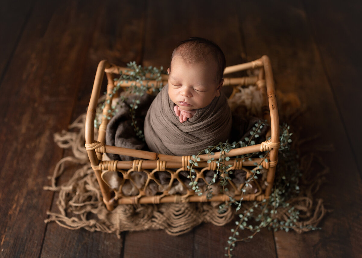 Newborn photoshoot | Baby wrapped in brown swaddle sitting in a newborn mini crib. Baby's hands are peeking out of swaddle under baby's chin.  Baby is sleeping.