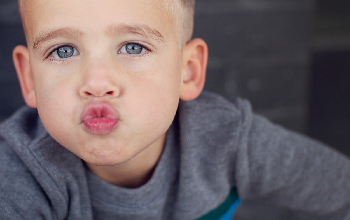 A little boy with blue eyes is making a kissy face at the camera on a gray background.