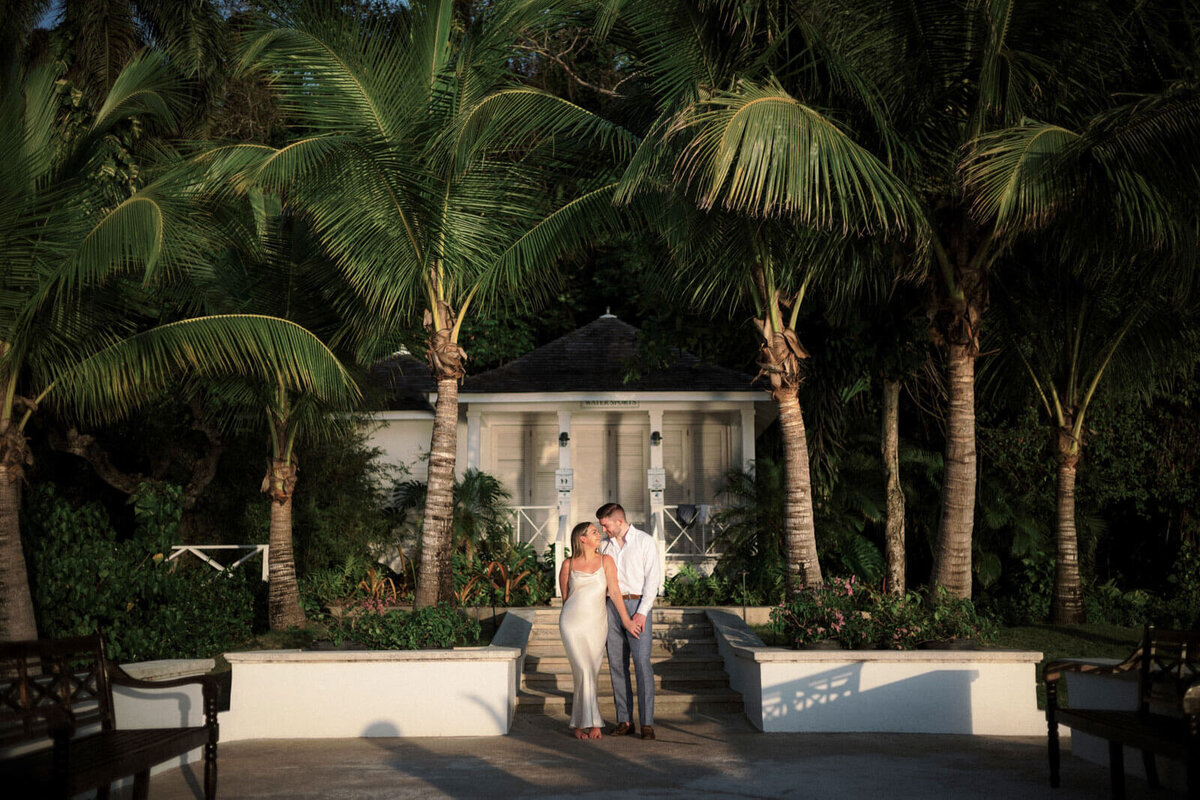 The engaged couple is standing in front of a villa, at Round Hill Hotel & Villas, Jamaica, with coconut trees in the background.