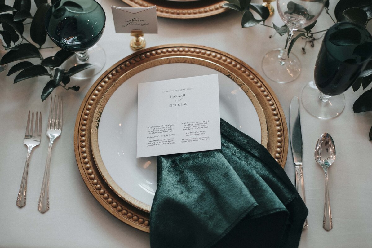 White squared place card with black font atop a dark green velvet napkin and white plate set on a reception table.