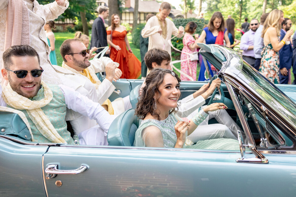 A joyful group of people dressed in festive clothing sits in a vintage convertible car at an outdoor event. The woman in front is smiling, and the men in the back seat are also cheerful. Other guests in colorful attire are visible in the background.