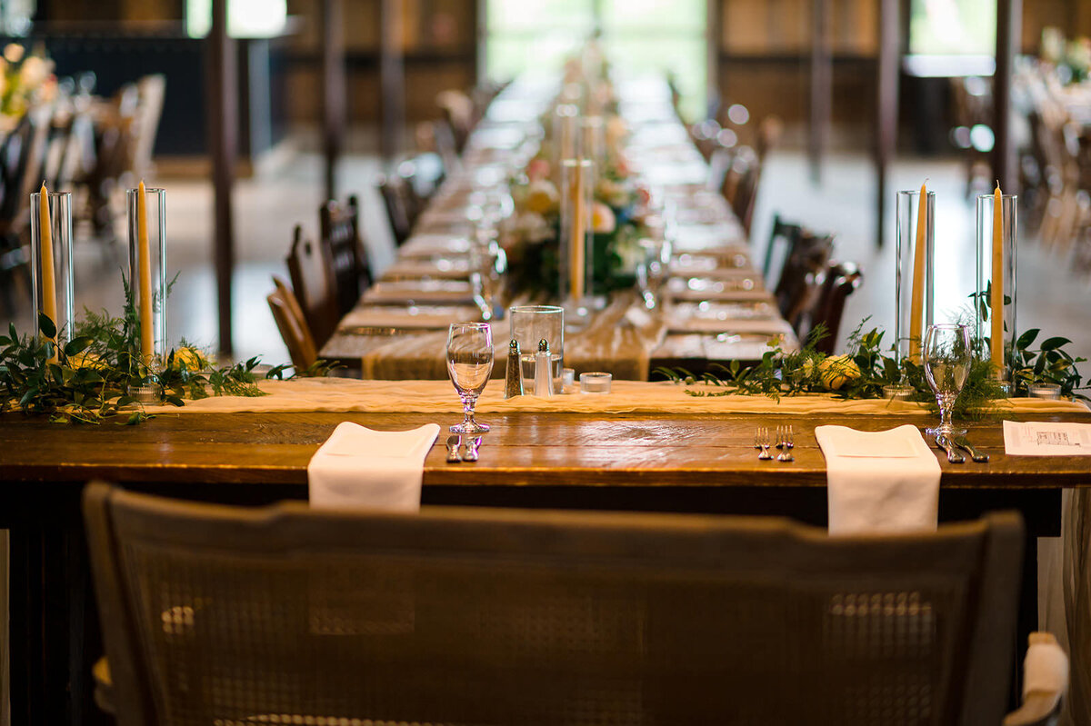 A long reception table set with greenery, candles, and place settings, ready for a wedding banquet in a dimly lit rustic venue.