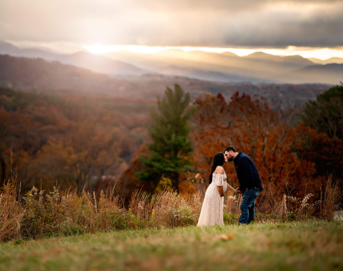 An adorable expecting couple smooch while standing in a field with the mounains behind them