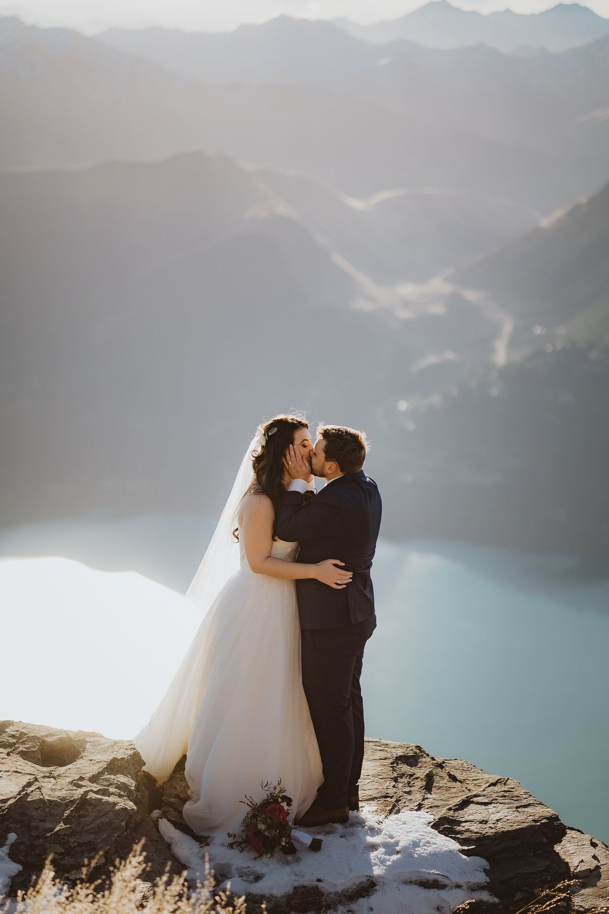 Groom grabbing his bride and kissing her on a cliff edge above a lake