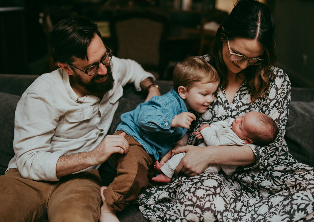 Playful family moment during newborn session—toddler reaching for baby sibling while parents laugh, captured in a cozy, in-home Portland shoot.