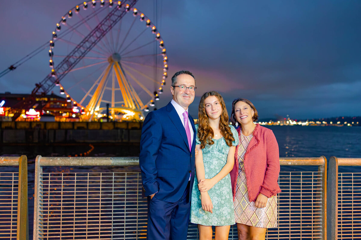 A mom and dad smile with their teenage daughter while standing on a waterfront walkway