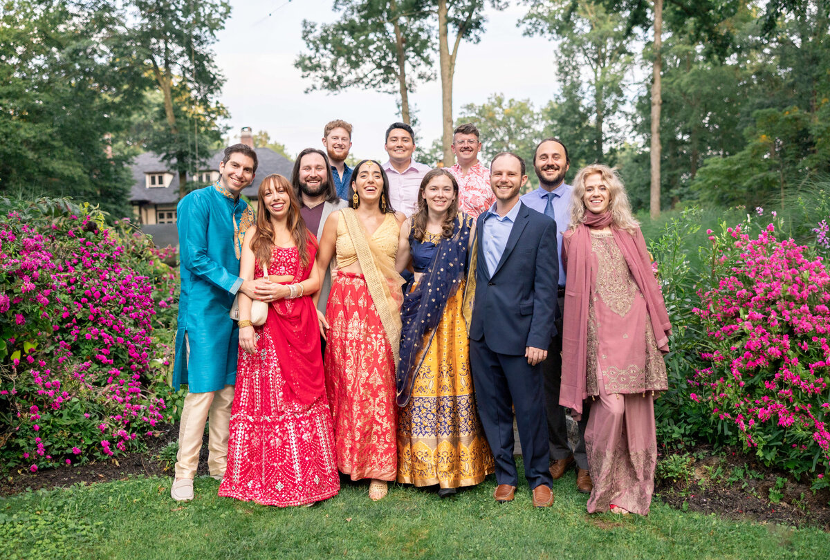 A group of people dressed in colorful traditional attire stand together outdoors, surrounded by blooming pink flowers and trees. They are smiling and posing for the photo on a lush green lawn.