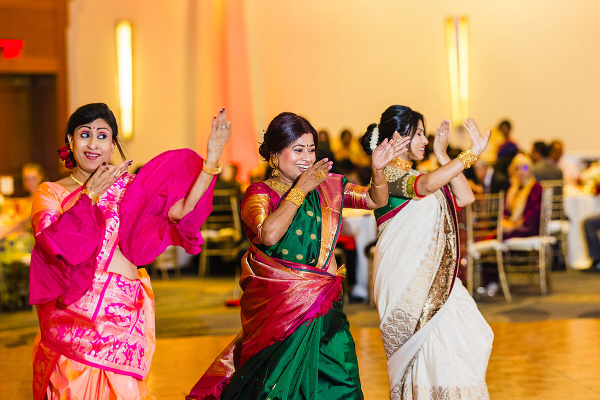 Three women in colorful sarees perform a traditional Indian dance with graceful hand movements at a wedding celebration.