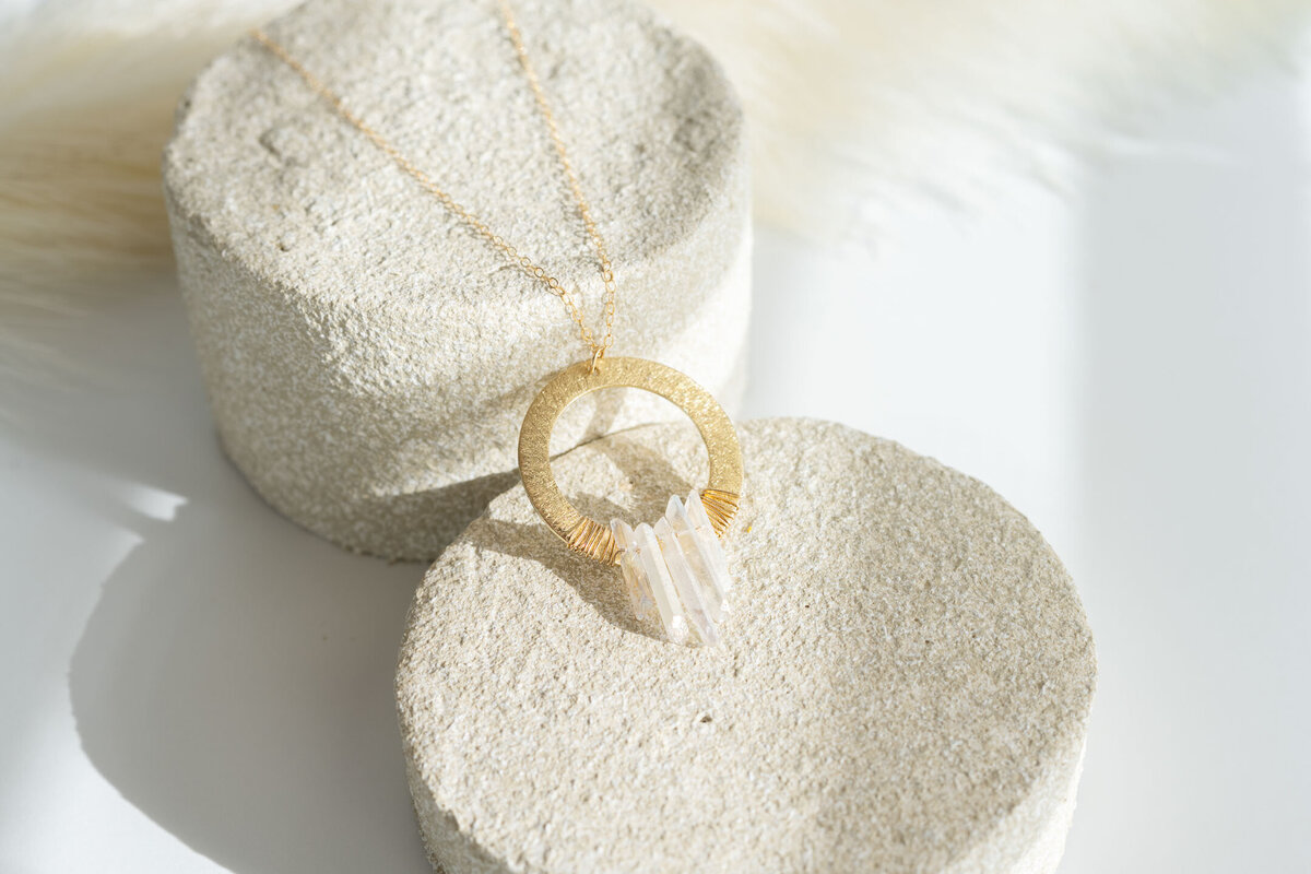 Gold necklace with a circular pendant and a crystal on round stone displays, bathed in soft sunlight.