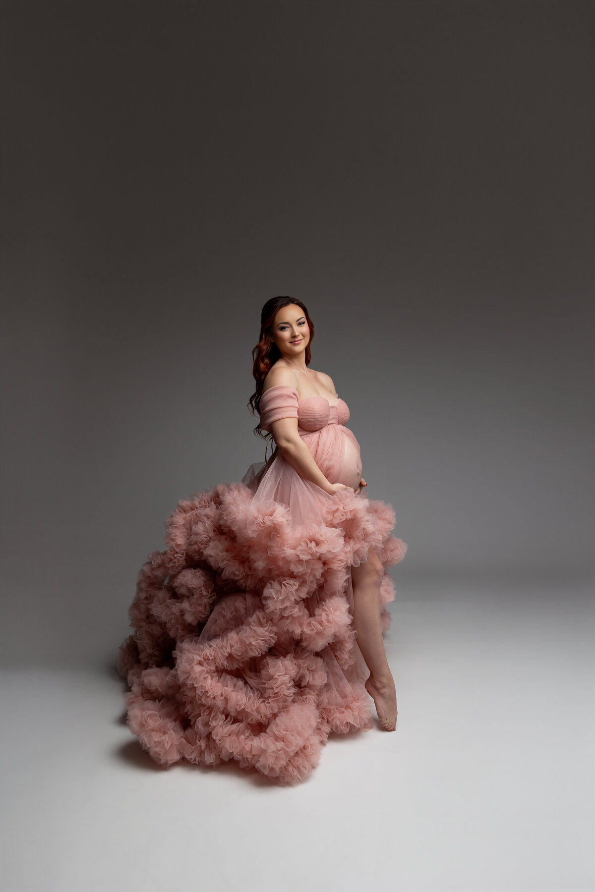 In a pink dress and poses in a side view, gently holding her belly with a serene expression. Captured by Inland Empire maternity photographer Kaitlyn Dawn Photography.