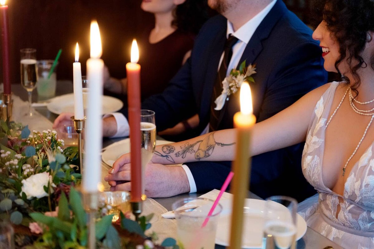 a bride and groom holds hands during wedding toasts.  you can see the bride's yellow bird tattoo between the glowing candles