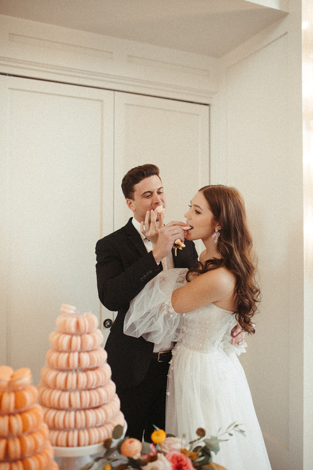 A bride and groom wearing a white wedding gown and black tuxedo feed macaroons to each other next to the dessert table.
