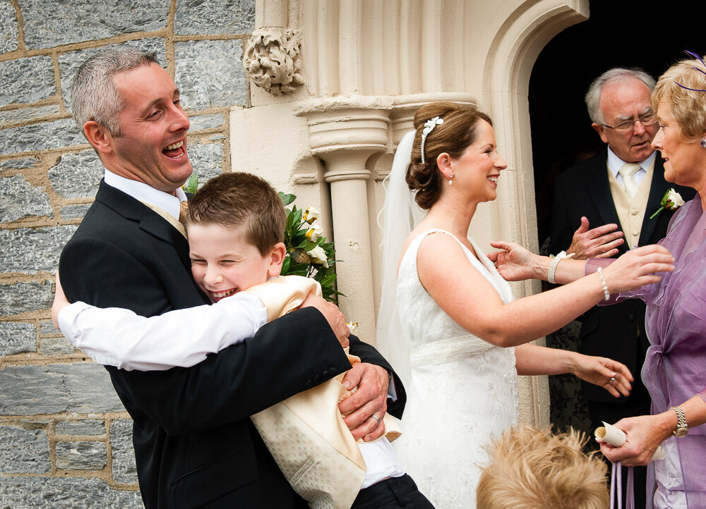happy groom being hugged by his page boy while laughing outside door of church in Kenmare