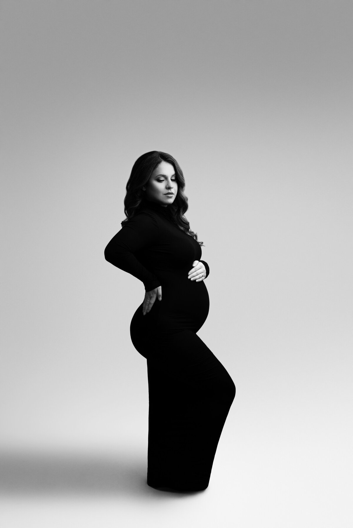 In this fine art maternity photo, Philadelphia Main Line's premier maternity photographer, Katie Marshall, captures a serene moment. The woman, dressed elegantly in a floor-length long-sleeve body-con dress, poses with her front leg gracefully bent. Her forehand cradles the small of her back, while her other hand gently rests atop her baby bump. With closed eyes, her face is angled tenderly toward her bump, creating a beautifully emotive and timeless composition