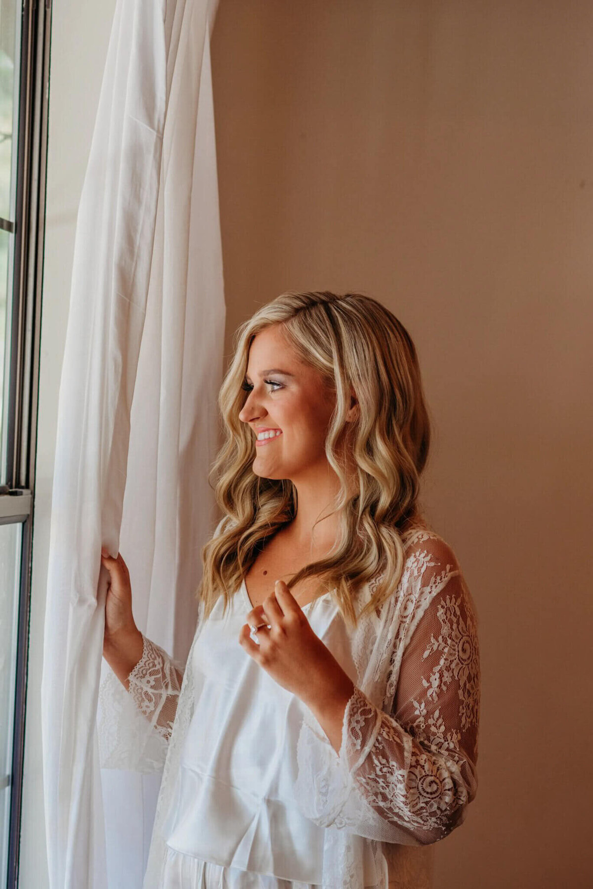 photo of a bride in a white robe while smiling out the window