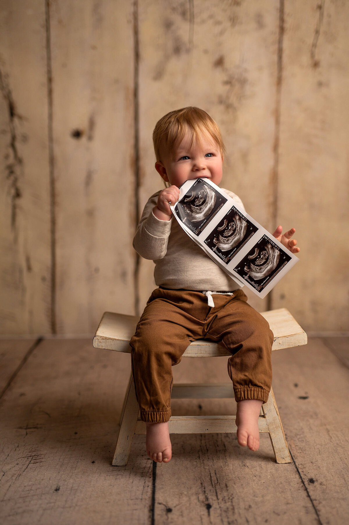 A toddler boy presents the ultrasound of his new sibling while seated on a stool in our bright Waukesha photo studio.