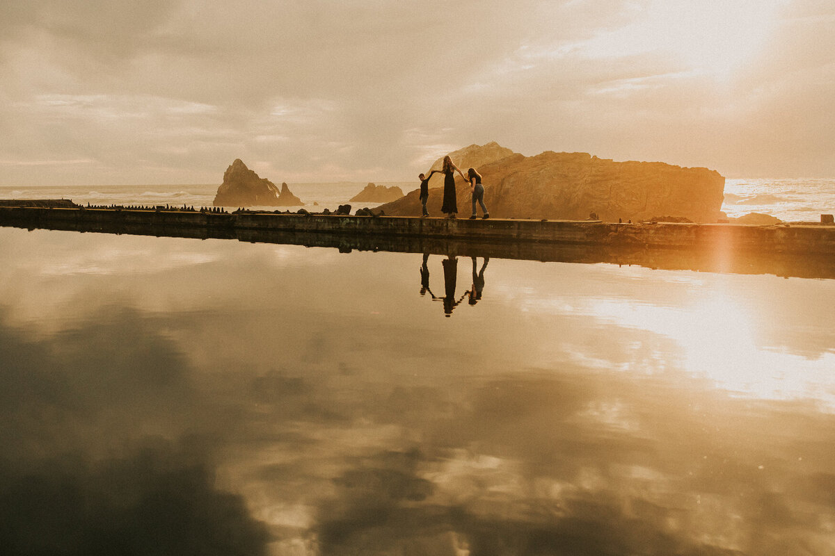 Wide angle documentary photo of mom with two children walking at Sutro Baths.  Warm sunlight and reflection of family on water