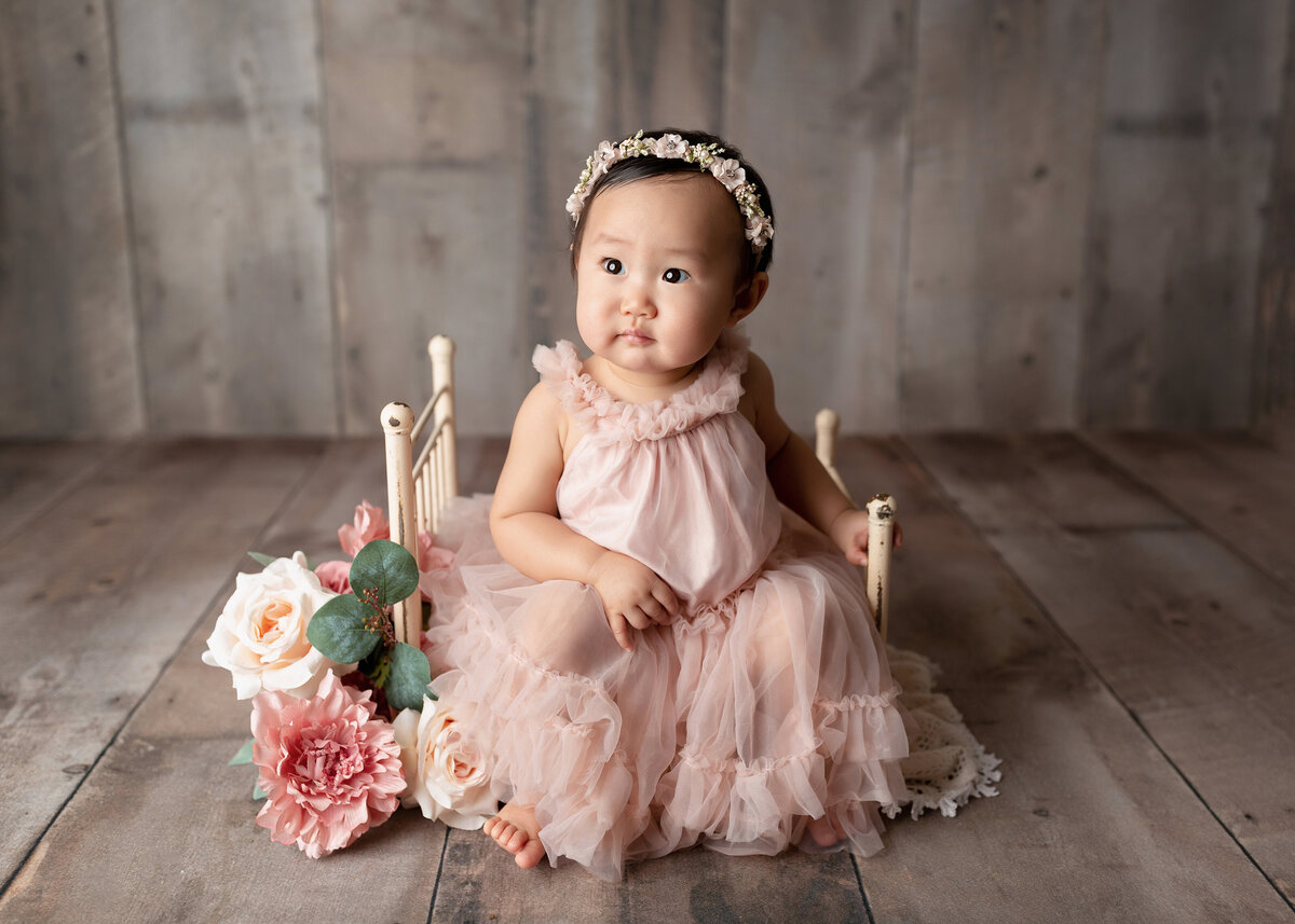12-month baby photoshoot at top  West Palm Beach and Boca Raton photography studio. Asian baby girl in a pink organza gown is sitting on a vintage metal doll bed. Baby is looking off in the distance.