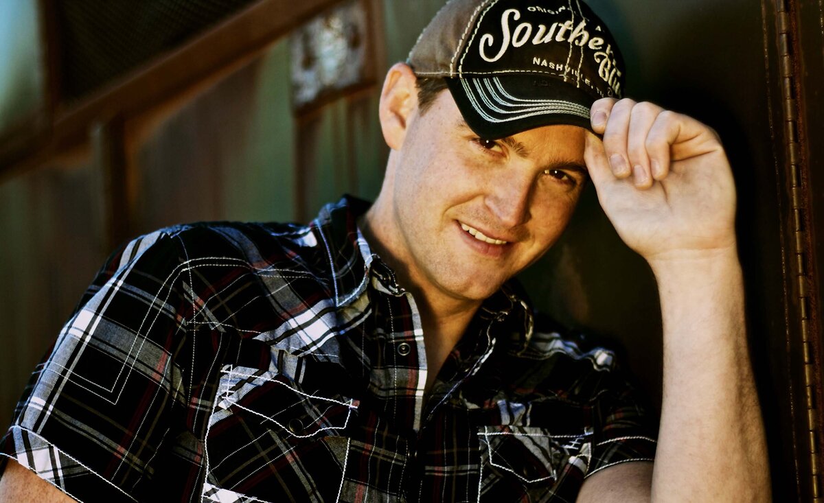 Country music photo Tristan Horncastle holding baseball cap brim leaning against green metal wall