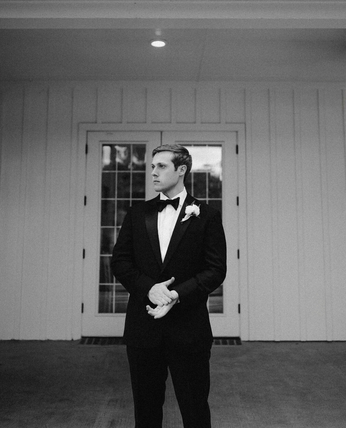 A man in a sleek black tuxedo and bow tie stands pensively in front of double glass doors. The monochrome tone adds a classic, timeless feel.