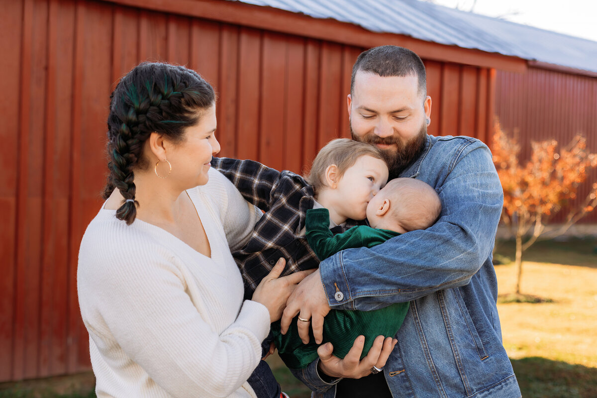 Family with New Baby and Toddler, standing in front of Red Barns for Updated Family Photos at Windsor Castle Park in Smithfield Virginia at Sunrise in Fall - Rebekah Heffington Photography